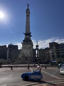 Aaron Novinger with a blue velomobile branded “PonziRelief.org” in front of the Soldiers and Sailors Monument at Monument Circle in Indianapolis, part of his cross-country Pedaling Against Ponzi’s charity ride from Dallas to Washington, D.C.