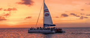 A catamaran named "Fury" sails on calm waters at sunset, with passengers enjoying the view against a vibrant orange and pink sky.