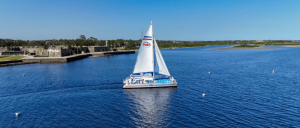 A white sailboat glides across calm blue waters near historic stone fortifications under a clear blue sky.