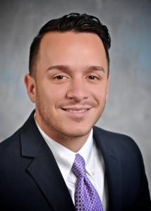 Portrait of a young man in a suit with a purple tie, smiling.