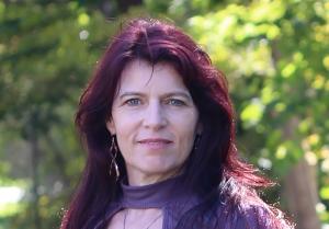 Headshot of Heather Abbott against a background of greenery. She has pale skin and dark red violet hair