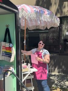 Founder/Owner of Beguiled Books, a white woman with brown hair, is smiling at the camera and placing a shopping bag on a hook