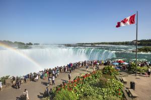 Crowds of visitors admire the Horseshoe Falls at Niagara Falls, Canada, on a sunny day, with a rainbow over the mist and a Canadian flag waving nearby.