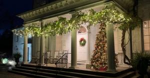 Front view of the mansion at Morven Museum & Garden in winter at night, decorated with white string lights, a Christmas tree, and a wreath on the front door.