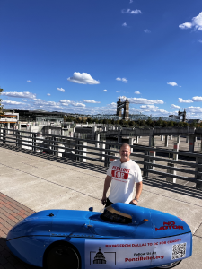 Aaron Novinger standing next to the John A. Roebling Suspension Bridge in Cincinnati next to his bright blue velomobile during his 2,000-mile charity ride, “Pedaling Against Ponzi’s,” to support victims of financial fraud.