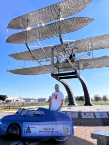 Aaron Novinger standing in front of the 1909 Wright Flyer airplane statue at Wright Patterson AFB next to his bright blue velomobile during his 2,000-mile charity ride, “Pedaling Against Ponzi’s,” to support victims of financial fraud.