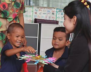 A Cambodian teacher helps two young students with a colorful hands-on classroom activity made from simple materials.