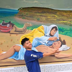 A scholarship student stands in front of a mural depicting the story of the Good Samaritan.