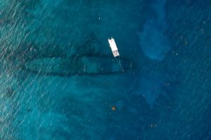 An aerial drone photo captures the clear, turquoise water above the SS Benwood shipwreck in Key Largo, Florida. The silhouette of the 300-foot-long WWII freighter wreck is visible beneath the surface. A 40-foot dive boat, the Siren, is moored directly ove