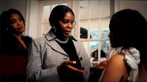 A dramatic still from The Caste of Beula trailer. A poised woman in a gray plaid blazer speaks intently to another woman in a white ruffled top, gesturing with her hands as tension builds between them. Another woman looks on from the background, adding to