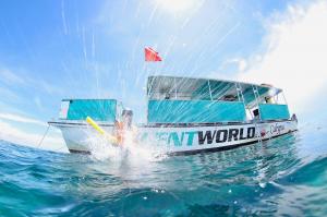 A point-of-view (POV) photo captures a snorkeler mid-jump, just after they've pushed off the back platform of a large boat. The snorkeler is framed against the bright turquoise, ultra-clear water, with a splash of white foam visible as they enter the wate