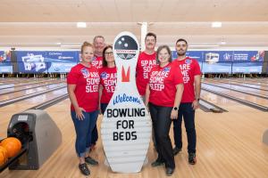 Six people wearing matching red t-shirts that read "Spare Some Sleep" pose with a large white bowling pin mascot at Victory Lanes bowling alley. The mascot has a smiling face and holds a "Welcome Bowling for Beds" sign. Bowling lanes are visible in the ba