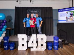 Three men stand on a stage holding a wooden award plaque. The man on the left wears a blue bowling shirt, the center recipient wears a red and white bowling shirt, and the man on the right wears a blue bowling shirt. Large white illuminated "B4B" letters