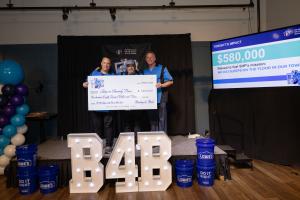 Three men in blue bowling shirts stand on a stage holding a large ceremonial check for $580,000 made out to Sleep in Heavenly Peace from Bowling for Beds. Large illuminated "B4B" letters are positioned in front of the stage, flanked by blue Lowe's buckets
