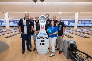 Seven people in matching black bowling shirts with red and white retro-style design stand with one person in a blue shirt behind a large white bowling pin mascot at Victory Lanes. The mascot displays a "Welcome Bowling for Beds" sign. The woman in blue ho