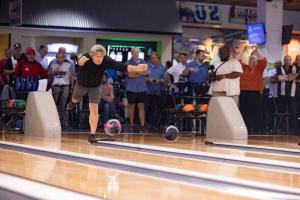 A bowler in a black shirt performs a dramatic trick shot with an exaggerated follow-through pose on a bowling lane. A crowd of spectators watches in the background, some recording with phones. Two bowling balls are visible on the lane behind him. The scen