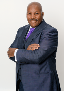 Dr. Lyman A. Montgomery standing confidently with arms crossed, wearing a navy suit, white shirt, and purple tie against a light background.