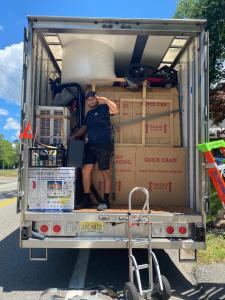 A Monroe Moving Pro employee stands inside a fully packed moving truck, smiling and pointing at the camera. The truck is neatly organized with labeled boxes, furniture, and moving supplies.