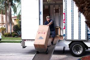 A Monroe Moving Pro mover rolls a large, fragile-labeled box down a ramp from a moving truck in a residential neighborhood. The mover appears focused and careful while unloading.