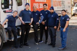 A group of six Monroe Moving Pro employees pose in front of a branded moving truck. They are dressed in uniform and smiling, showing team spirit and professionalism.