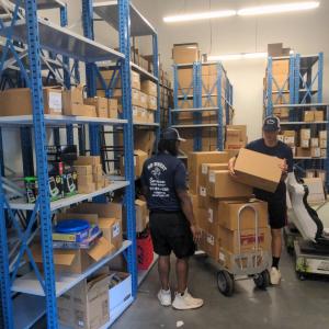Two movers from No Sweat Services Inc. work in a warehouse, loading and organizing boxes onto a hand truck between blue metal shelving units filled with packages.