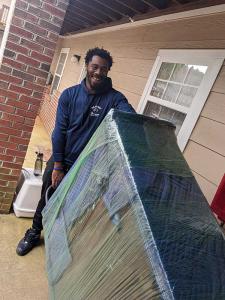 A smiling No Sweat Services Inc. mover stands outside a home with a large furniture item wrapped in moving blankets and plastic wrap on a dolly.