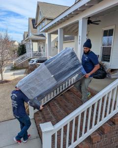 Two No Sweat Services Inc. movers carry a large, shrink-wrapped piece of furniture up a brick staircase into a home. One mover is lifting from the bottom, and the other guides from the top.