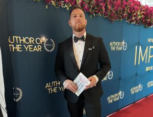 Joshua Atlas Aultman stands in a black tuxedo with a pirate themed bow-tie holding his book Rule of 3 Leadership - How Elite Leaders Win while standing on the red carpet in the Hollywood Bowl area in front of a step and take banner with logos that read “A