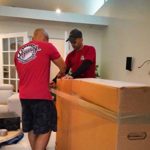 Two movers from Skyway Moving & Storage are sealing a large cardboard box in a living room, demonstrating careful packing techniques.