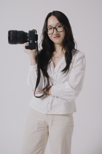 Audrey Tjahjono holding a camera, wearing glasses, a white long-sleeved shirt, and matching white pants. Audrey has long hair down and stands confidently, ready to capture photos.