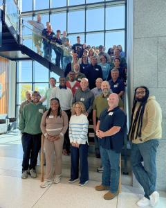 A diverse group of approximately 40-50 Sleep in Heavenly Peace volunteers posed together on and around a modern staircase in a bright building with floor-to-ceiling windows. The group includes men and women of various ages wearing casual clothing and name