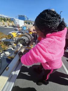A woman in a pink fuzzy sweater uses a DEWALT power saw on a piece of lumber while being guided by an instructor in a red SHP polo shirt. They are working at an outdoor training workshop with other volunteers visible in the background. The setting appears