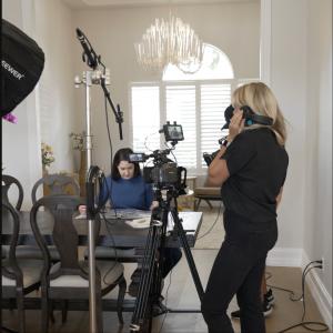 Director Rebecca Hodges stands on set, monitoring audio with one headphone to her ear, while Executive Producer and protagonist Elizabeth Forrest sits at a table flipping through a book. Studio lights and cameras focus on her during the filming of “Beyond