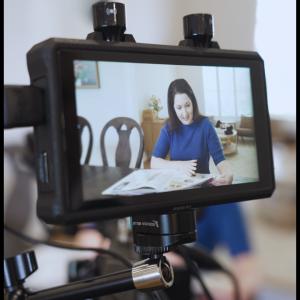 A close-up of a camera preview monitor showing Executive Producer and protagonist Elizabeth Forrest smiling and speaking as she flips through a scrapbook. The monitor display captures her in soft lighting during the filming of “Beyond the Diagnosis.”