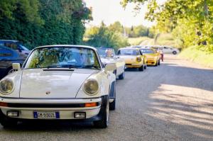 Vintage car parade arriving at Santa Giustina Abbey in Piedmont, classic Porsche and Ferrari convoy.