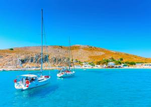 Two serene sailboats glide across the azure waters of Greece, passing by a hidden gem of an island, perfectly capturing the essence of authentic travel experiences by Tourmix.tours – with our unmatched know-how, we take you where others can only dream, so