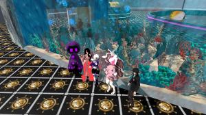 A vibrant group of five colorful avatars pose inside a virtual reality aquarium lounge at the Ferry Godmother Roller Rink. The scene features a large curved glass tank filled with coral reefs, tropical fish, and glowing blue bubbles. The avatars stand on