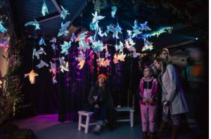 A group of visitors stands and sits beneath dozens of glowing paper birds suspended from the ceiling in a dimly lit room. The scene is bathed in soft purple, blue, and orange light, creating a serene and contemplative atmosphere.