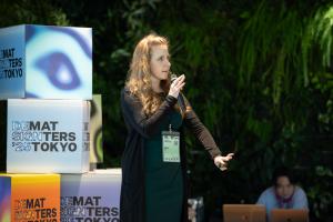 Cheryl Platz, a professional blonde woman wearing a green dress and black jacket, stands on a modern-looking stage surrounded by plants, computers, and colorful blocks while giving a talk.