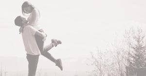 A black-and-white image of a young man lifting a smiling young woman in his arms outdoors; she has one leg kicked up, and they share an affectionate gaze amid a background of bare trees and hazy sky.