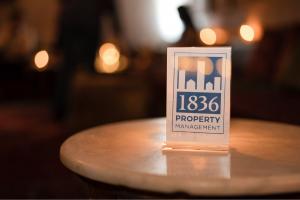 Clear plastic stand holding a card with 1836 Property Management logo on a light round table with blurred warm lights and reflections.