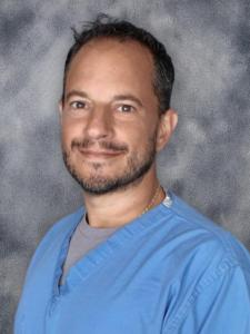 Dr. Nicholas Verdura, MD, FACS, CWS-P, newly appointed Medical Director of Advanced Wound Care and General Surgery at Confidia Health Institute in Plantsville, Connecticut, wearing blue medical scrubs and smiling against a gray background.