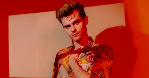 A young man, Theo Walker, who portrays Sebastian in the film Nosebleed, stands against a warm orange background. There are hues of hot reds that slice across his sharp features. He is wearing a funky-patterned short sleeved shirt with the top buttons undo