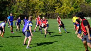 Rugby players wearing red and blue vests in mid-play on a sunny day, as one player runs with the ball while others prepare to defend, with trees showing early autumn colours in the background.
