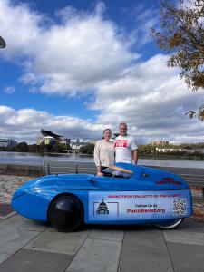 The Novinger's standing in front of the Acrisure Stadium in Pittsburgh next to the bright blue velomobile during their 2,000-mile charity ride, “Pedaling Against Ponzi’s,” to support victims of financial fraud.