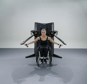 A person seated in a wheelchair performing a cable fly exercise using a dual-adjustable pulley. The individual’s arms are extended outward, holding the machine’s handles, with the equipment positioned directly behind her.