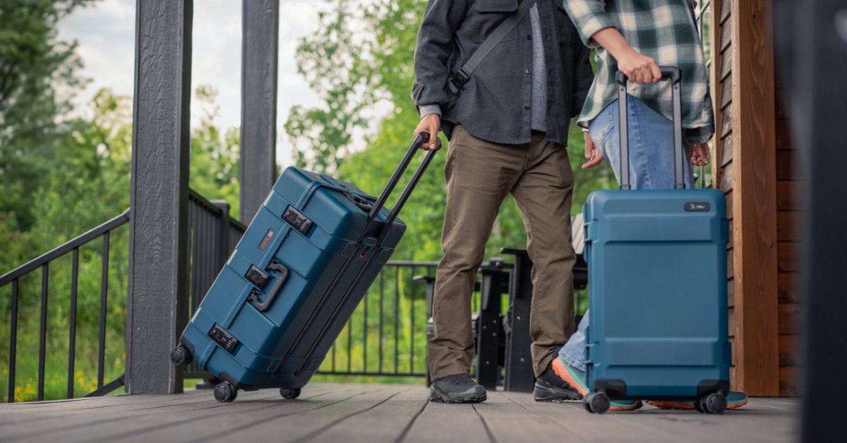 Two travelers pulling rugged blue Peli hard-shell luggage with wheels on a wooden porch, surrounded by green outdoor scenery, showcasing durable and stylish travel protection.