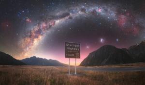 A star-filled sky and Milky Way are seen above a mountain landscape at night with a road sign that reads Starlight Highway.