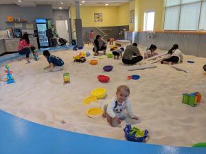A bright indoor sand play space with children and adults playing with sand toys, buckets, and tubes at Sandmagination.