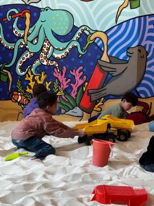 Children playing with sand toys in a bright indoor sandbox space with a large ocean mural behind them.